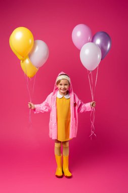 Uzun boylu, yağmurluklu, kauçuk çizmeli, kırmızı balon tutan mutlu bir kız.