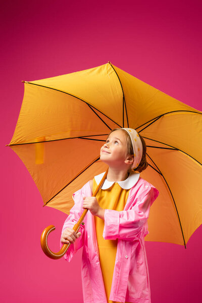 pleased girl in raincoat standing under yellow umbrella isolated on crimson