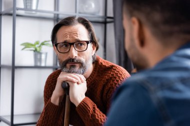 Elinde baston olan üzgün adam bulanık ön planda oğluna bakıyor.