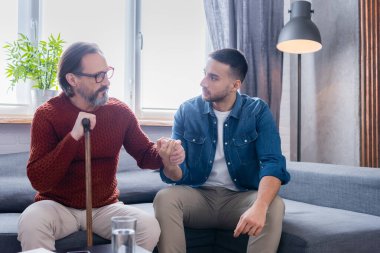 Elinde baston olan olgun bir adam koltukta otururken genç İspanyol oğluyla el ele tutuşuyor.