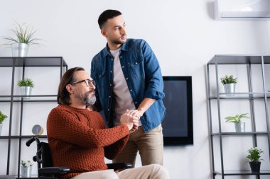 Engelli bir adam tekerlekli sandalyede oturmuş gözlerini kaçıran İspanyol bir çocuğun elini tutuyor.