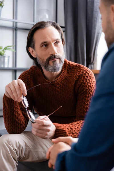 Bulanık ön planda oğlunu dinlerken gözlük tutan olgun bir adam.