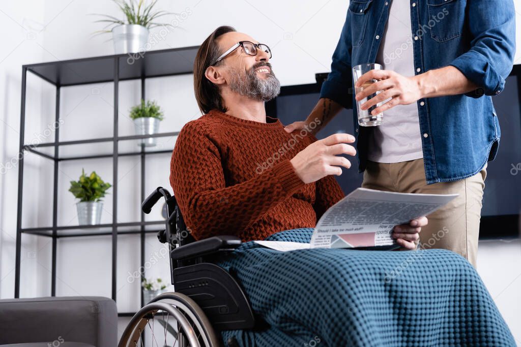 young man giving glass of water to smiling handicapped man sitting with newspaper in wheelchair