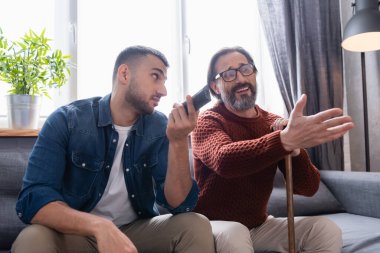 Elinde uzaktan kumandayla oğlunun yanında televizyon seyreden hoşnutsuz bir adam.