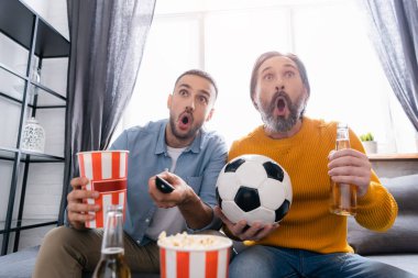 Irklar arası baba-oğul, bira ve patlamış mısırın yanında futbol maçı izliyorlar.