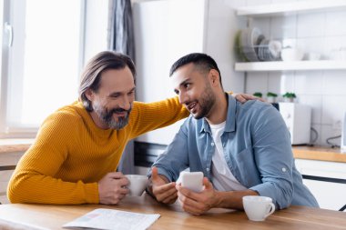 Gülümseyen adam, İspanyol oğlunu kucaklayarak mutfaktaki akıllı telefonu işaret ediyor.