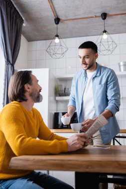 Mutfakta babamın yanında kahve fincanı tutan genç İspanyol adam, bulanık ön plan.