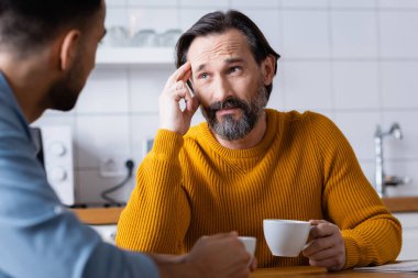Sakallı bir adam oğluyla mutfakta konuşurken kafasına dokunuyor, ön planı bulanık.