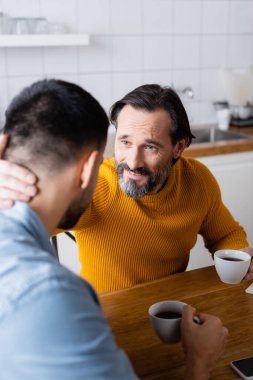 Gülümseyen bir adam mutfakta konuşurken boynuna sarılıyor, ön plan bulanık.