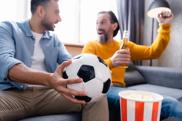 young hispanic man with soccer ball near excited father showing success gesture, blurred background