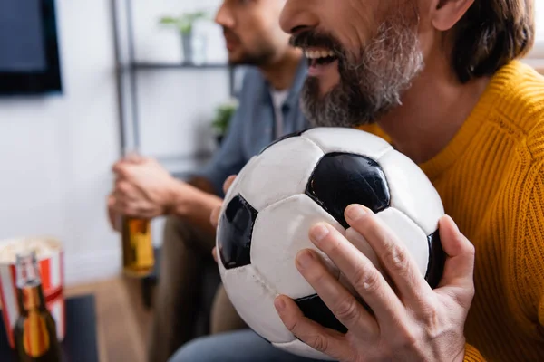 cropped view of excited man holding soccer ball while watching championship together with son on blurred background