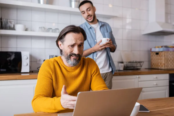 Skeptical Man Pointing Laptop Smiling Hispanic Son Cup Blurred Background — Stock Photo, Image