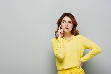 pensive woman biting lip while standing with hand on hip on grey clipart