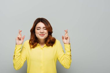 positive woman holding crossed fingers while standing with closed eyes on grey clipart