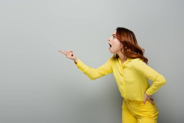 shocked woman pointing with finger while standing with hand on hip on grey clipart
