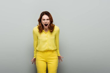 angry woman in yellow clothes yelling while looking at camera on grey clipart