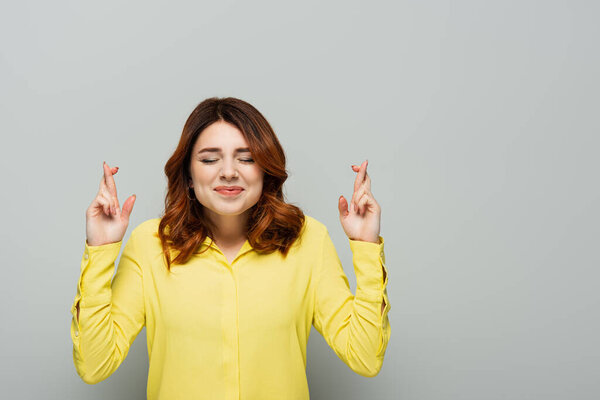 positive woman holding crossed fingers while standing with closed eyes on grey