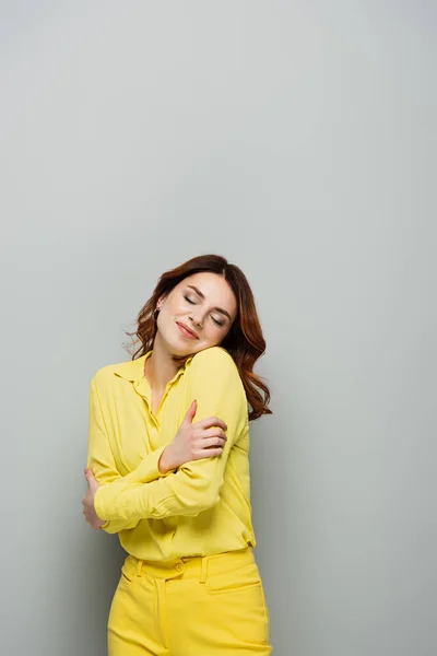 pleased, dreamy woman hugging herself while standing with closed eyes on grey