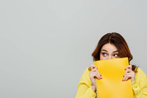 испуганная женщина смотрит в сторону, скрывая лицо с блокнотом, изолированным на серой