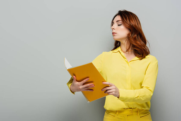 smug woman in yellow shirt looking at notebook while standing on grey