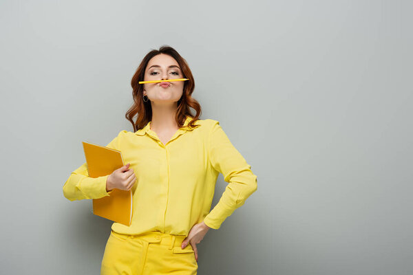 кудрявая женщина с карандашом между носом и губами, позирующая рукой на бедре на сером