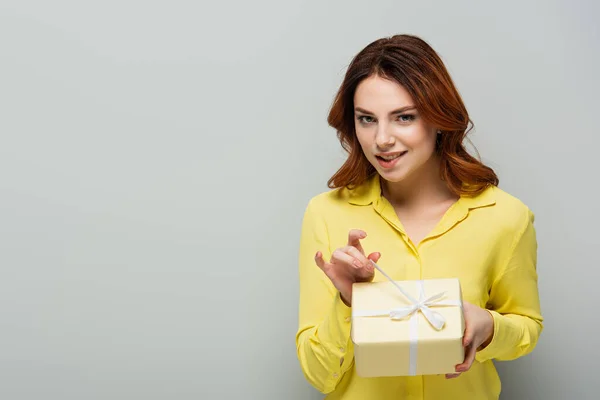 flirty woman opening gift box while looking at camera on grey