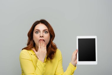 Gri ekranda izole edilmiş boş ekranlı dijital tableti tutarken ağzı eliyle kapatan şok olmuş bir kadın.