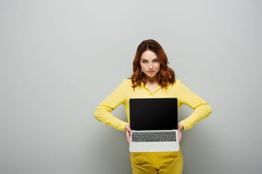 smirking woman looking at camera while holding laptop with blank screen on grey clipart