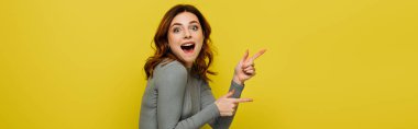 excited woman looking at camera while pointing with fingers isolated on yellow, banner clipart