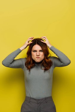 Kıvırcık saçlı stresli kadın kafasına dokunuyor ve sarı kameraya bakıyor.