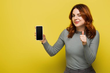 Kıvırcık saçlı neşeli kadın elinde akıllı telefon, ekranı boş ve sarı başparmağını gösteriyor.