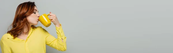 curly woman in yellow blouse drinking tea on grey, banner