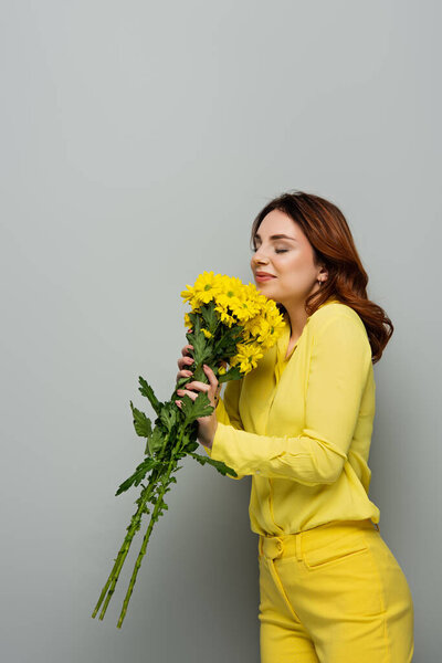 pleased woman holding yellow chrysanthemums while standing with closed eyes on grey