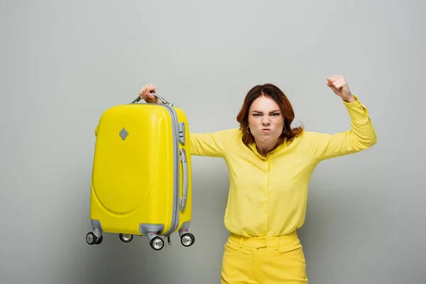angry woman showing clenched fist while holding yellow suitcase on grey