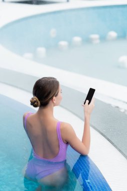 Açık havuzda yüzerken elinde boş ekranla akıllı telefon tutan genç bir kadın.