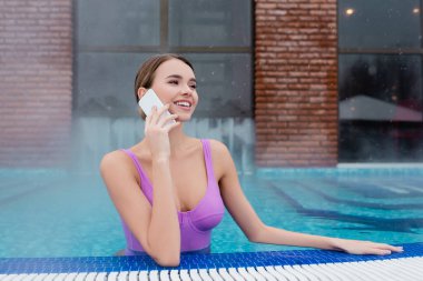 smiling woman in swimsuit talking on mobile phone while bathing in outdoor swimming pool clipart