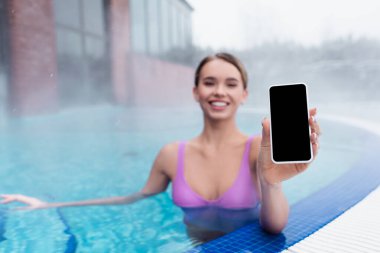 Bulanık ve neşeli bir kadın açık havuzda banyo yaparken elinde boş ekranla akıllı telefonu tutuyor.