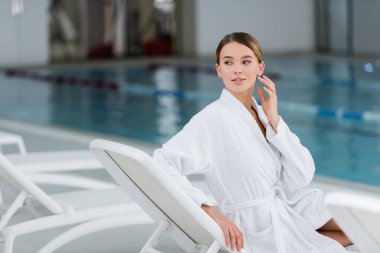 Beyaz bornozlu genç bir kadın havuz kenarındaki iskemlede dinlenirken gözlerini kaçırıyor.