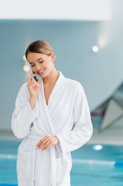 Beyaz bornozlu mutlu genç kadın spa merkezinde akıllı telefondan konuşuyor. 
