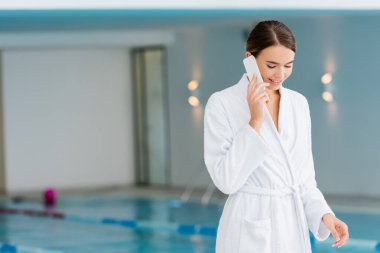 Beyaz bornozlu neşeli genç kadın spa merkezinde akıllı telefondan konuşuyor. 