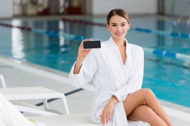 Beyaz bornozlu genç bir kadın elinde beyaz ekranlı akıllı bir telefon tutuyor. 