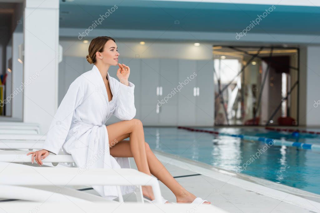 happy young woman in bathrobe sitting on deck chair near pool in spa center 