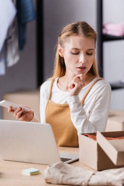 Düşünceli bir galeri sahibi iş yerinde dizüstü bilgisayar ve kartonun yanında akıllı telefon tutuyor.
