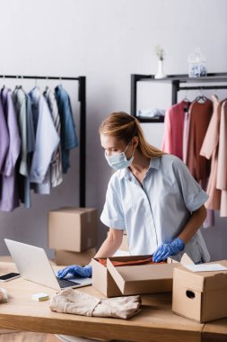 Tıbbi maskeli genç bir iş kadını, galeride dizüstü bilgisayarın yanında sipariş topluyor.