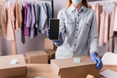 Ön planda bulanık kutuların yanında akıllı telefon tutan lateks eldivenli sergi sahibinin kırpılmış görüntüsü