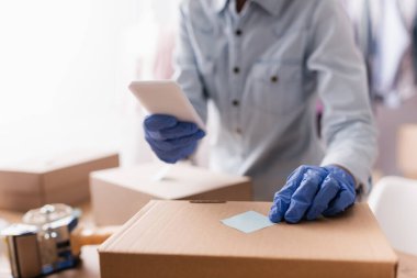 Lastik eldivenli satıcının elinde akıllı telefon, bulanık arkaplan tutarken kutudaki yapışkan notları düzelttiği kırpılmış görünüm