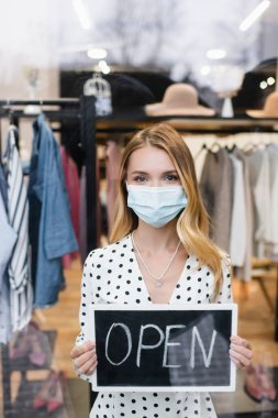 Tıbbi maskeli pozitif bir iş kadını. Moda mağazasında açık harflerle pano tutuyor.