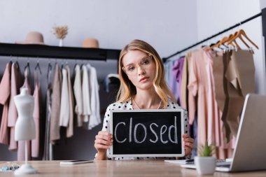 upset businesswoman holding board with closed lettering in showroom, blurred foreground clipart