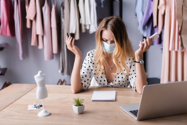Tıbbi maskeli kaşları çatık bir iş kadını, elinde akıllı telefon ve kalemle sergi odasında boş defterin yanında.