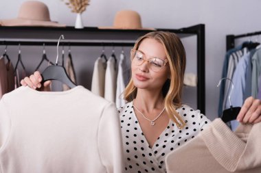 joyful successful showroom owner holding hangers with clothes on blurred foreground clipart